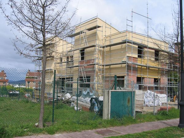 Edificio en construcción, fachada beige, andamios, valla verde, día nublado.