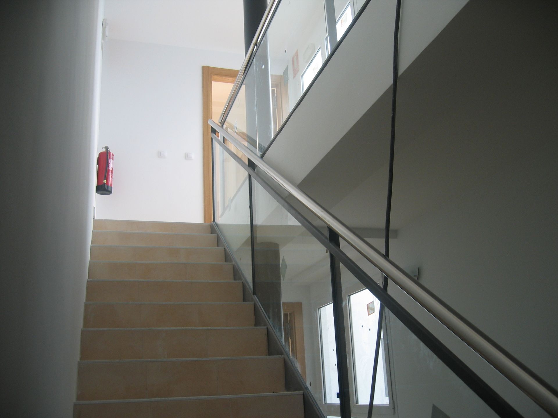 Escalera con barandilla de cristal que sube. Extintor en la pared de la izquierda.