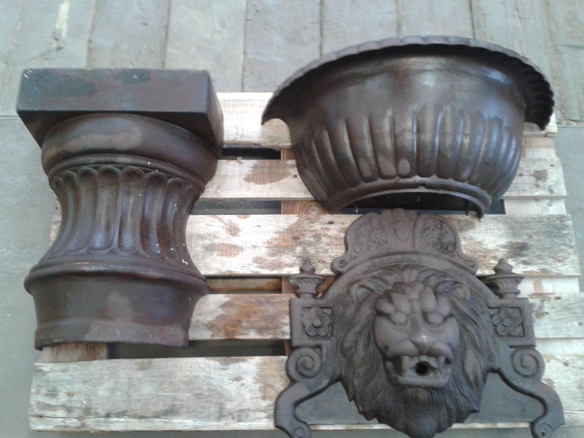 Éléments décoratifs en fonte : un socle de colonne, une jardinière et une tête de lion, sur une palette en bois.