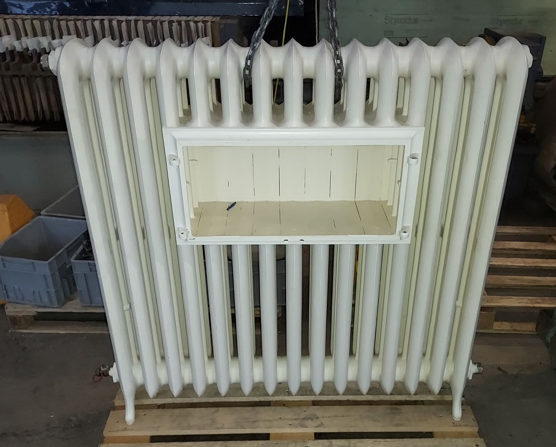 Grand radiateur blanc cassé avec étagères rectangulaires intégrées. Il repose sur une palette en bois dans un atelier.