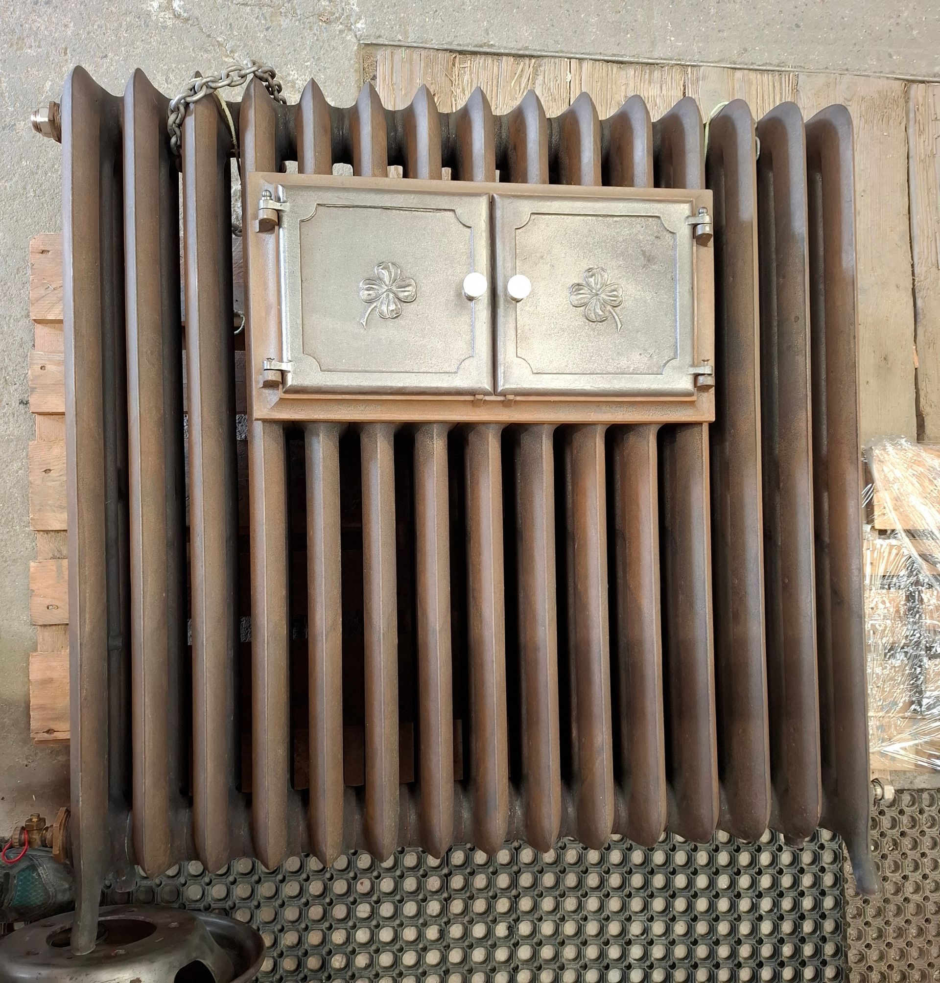 Un vieux radiateur à vapeur en fonte, doté d'un petit panneau d'accès à double porte. De couleur brune, il se détache sur un fond en bois.