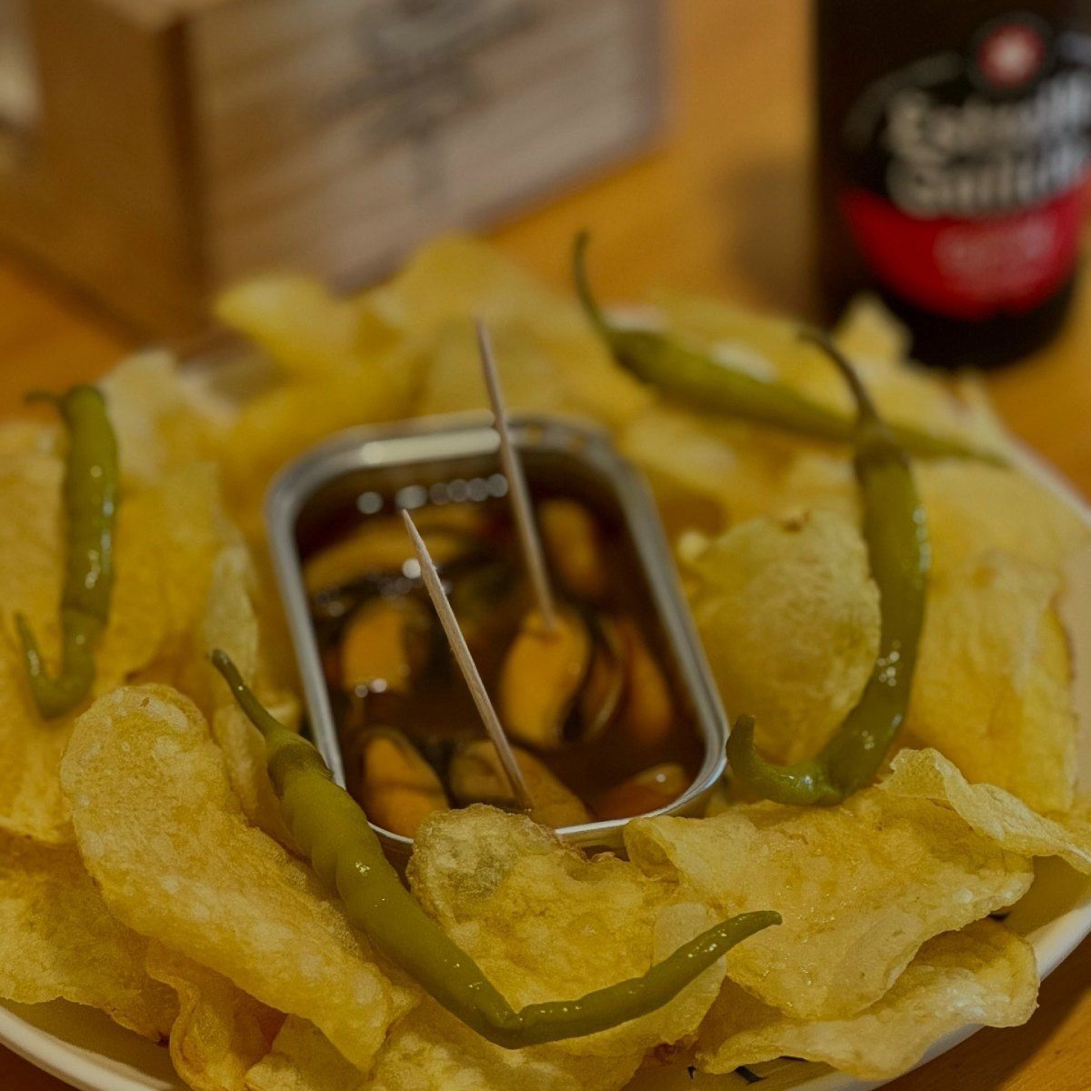 Plato de patatas fritas, mejillones enlatados en salsa y pimientos verdes, con una botella de cerveza de fondo.