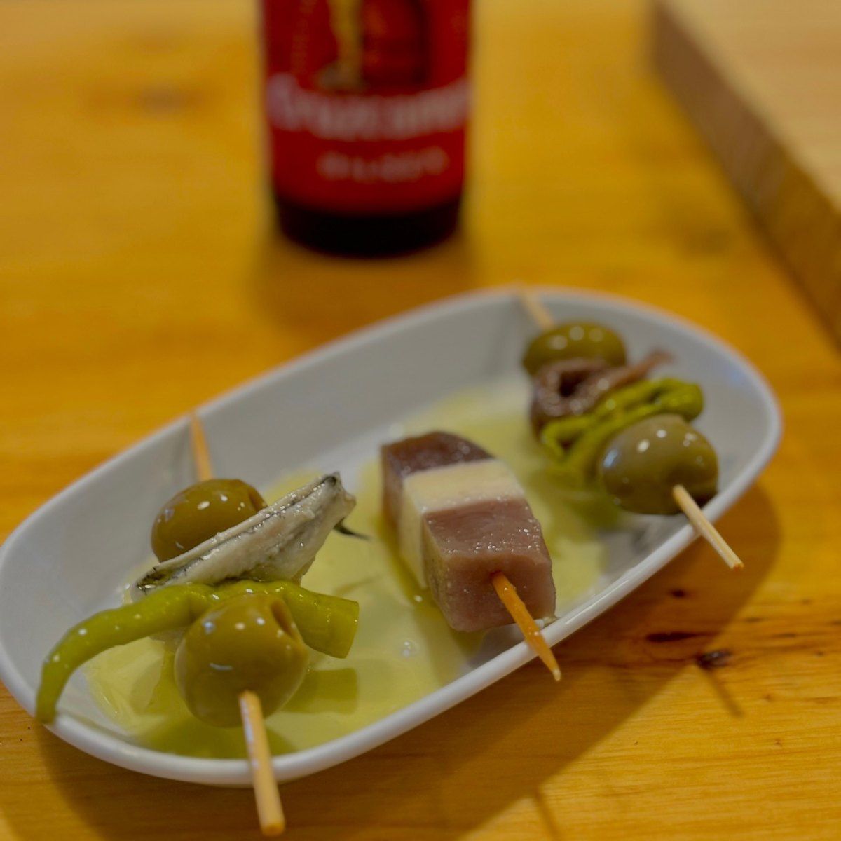 Pequeñas brochetas de tapas en plato blanco, con una botella de cerveza de fondo.