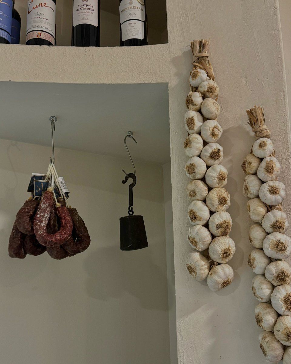 Salchicha y peso colgando de ganchos, junto a ristras de ajos. Botellas de vino en el estante superior.