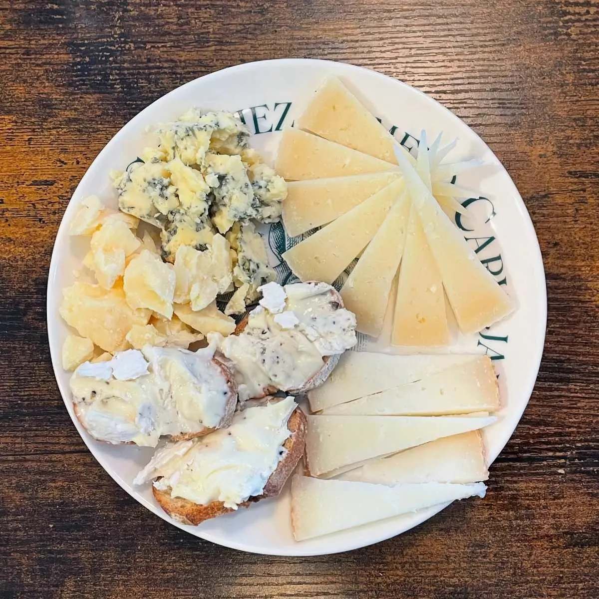 Tabla de quesos: queso azul, queso curado en lonchas, pan cubierto de brie y otros quesos en un plato blanco.