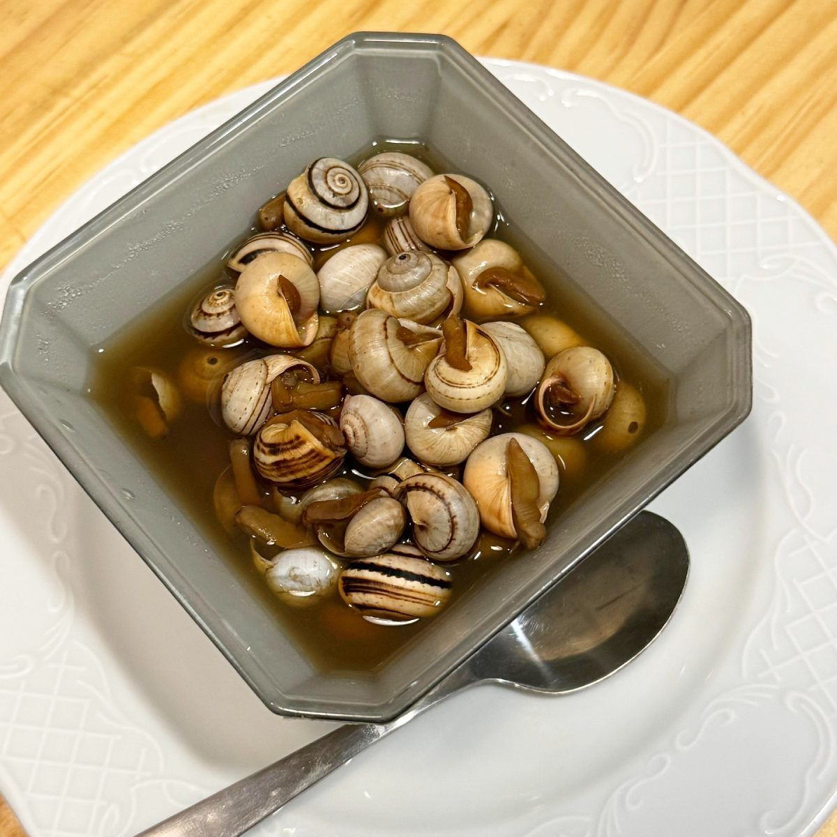 Cuenco de caracoles cocidos en caldo, en un plato blanco con una cuchara.