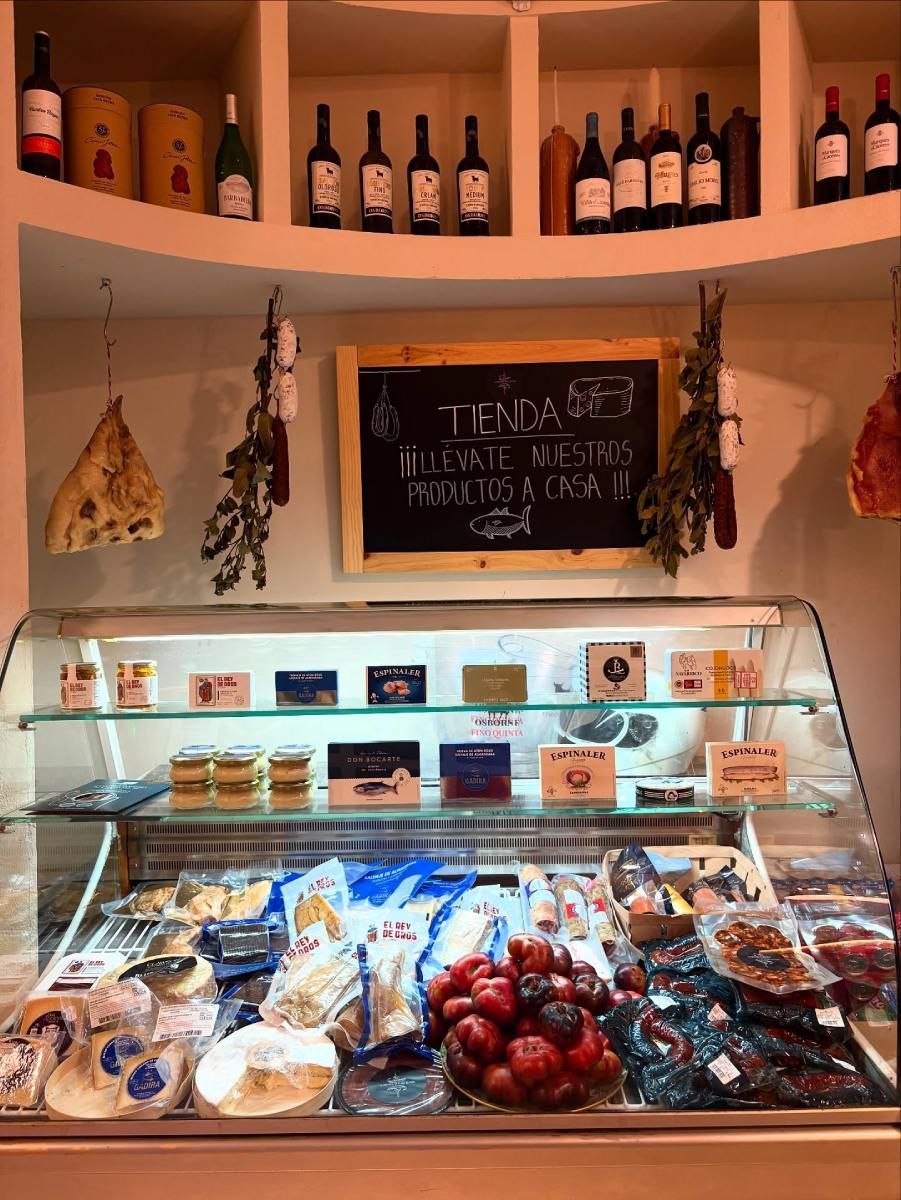 Vitrina de delicatessen con diversos alimentos, botellas de vino en el estante superior y cartel de pizarra.