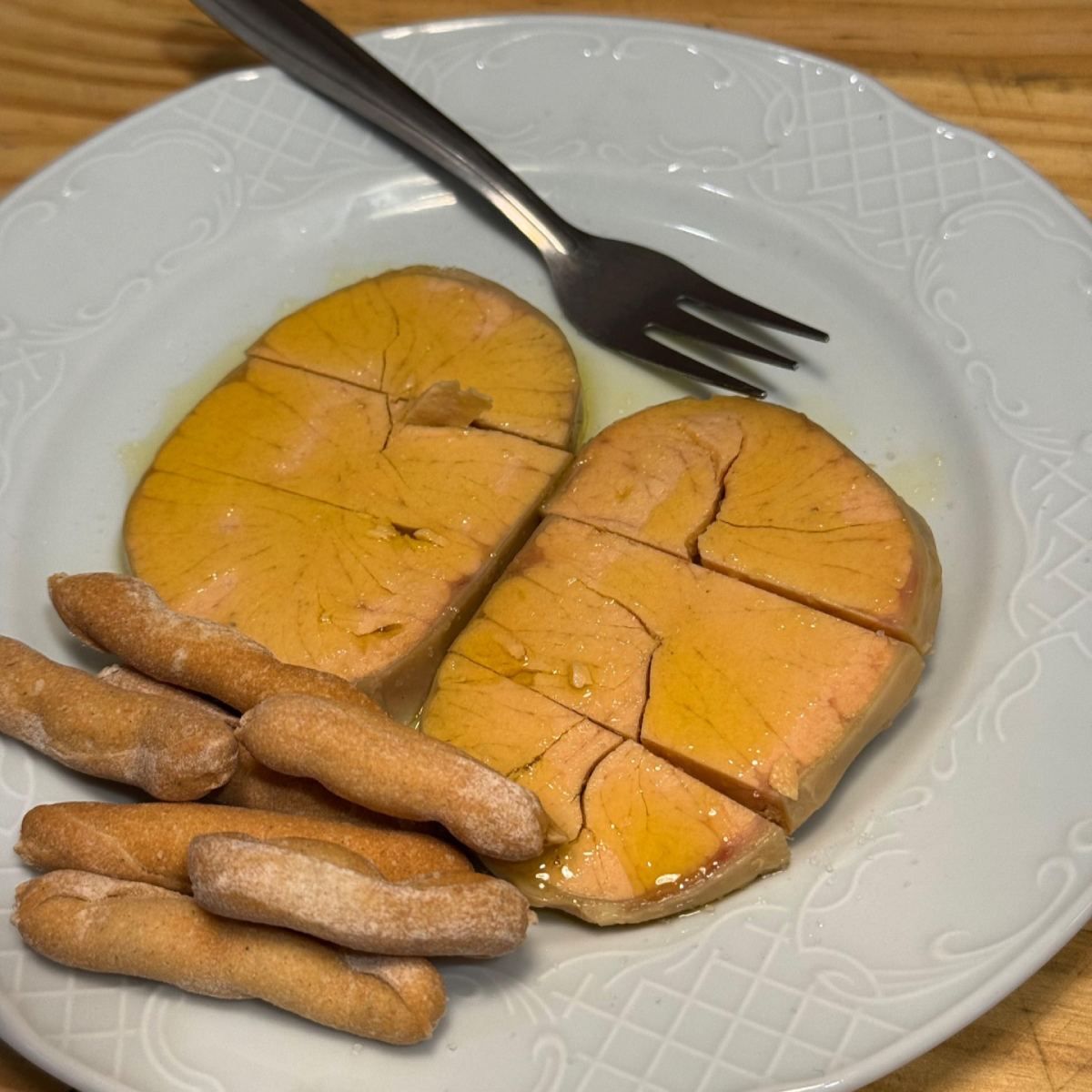 Plato con dos porciones de foie gras cortado en cuadritos y sellado en dorado, palitos de pan y un tenedor.