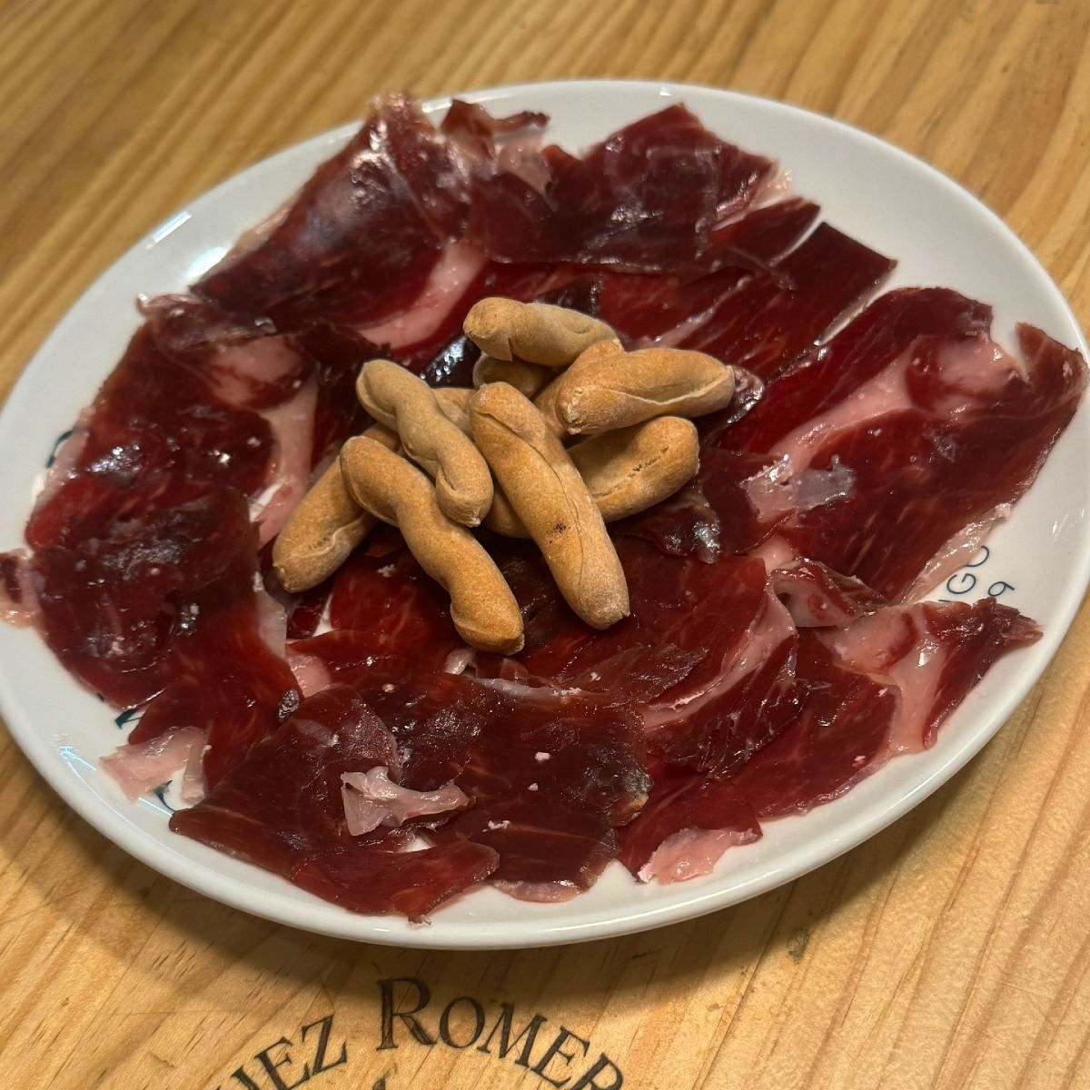 Plato de jamón curado cortado fino y palitos de pan sobre una superficie de madera.