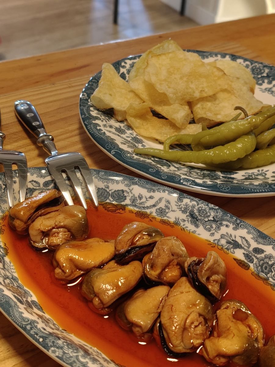 Mejillones en salsa en un plato ovalado, con patatas fritas y pimientos verdes en un plato aparte, sobre una mesa de madera.