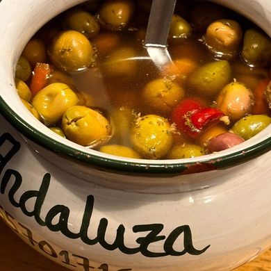 Una olla de cerámica con aceitunas en salmuera con un cucharón; en la olla se puede leer "Andaluza".