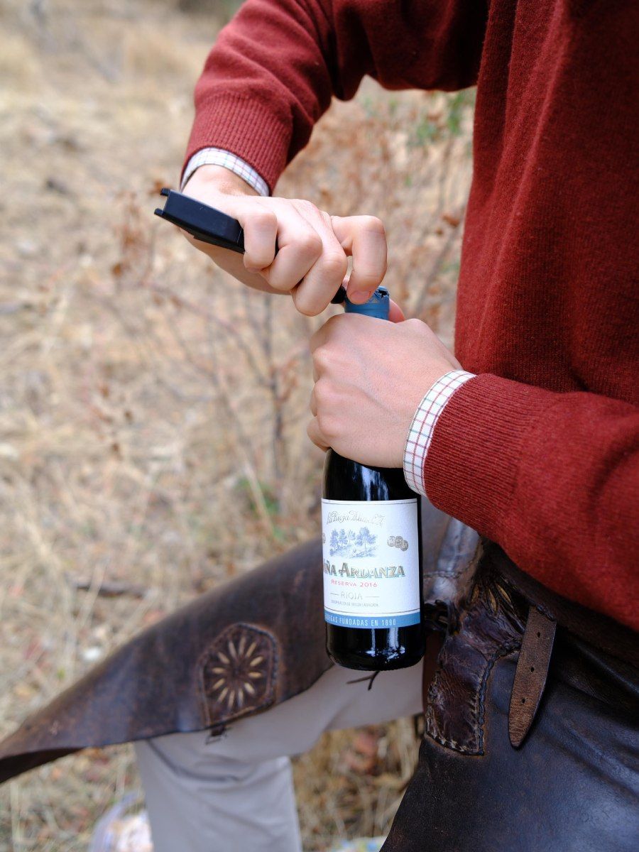 Hombre con suéter rojo abriendo una botella de vino con un sacacorchos negro al aire libre, vistiendo polainas.