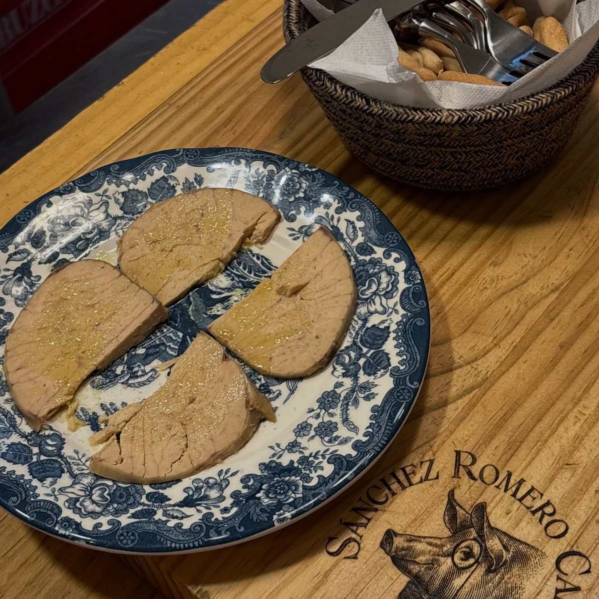 Plato de carne cortada en rodajas con aceite sobre un plato con estampado azul y blanco, cesta de pan cerca.