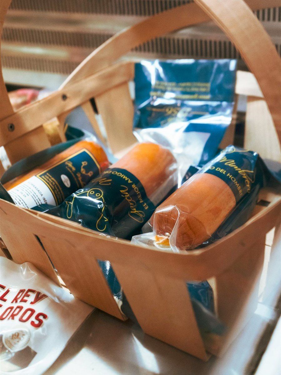 Una cesta que contiene ruedas de queso envueltas y otros productos envasados, en una cesta de madera.