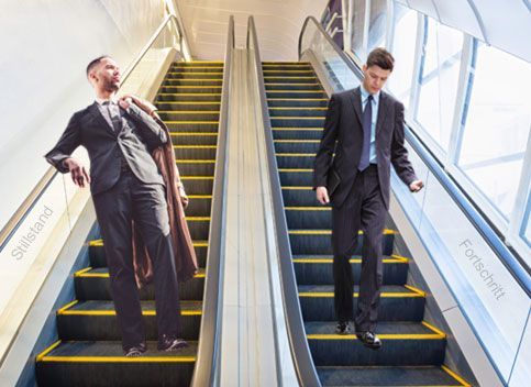 Zwei Männer stehen auf zwei Rolltreppen die nach unten fahren