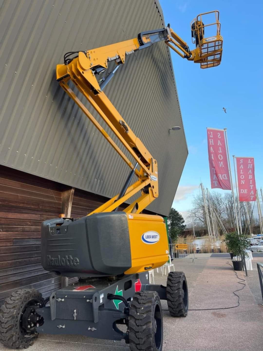 Nacelle élévatrice JLG jaune à côté d'un bâtiment, bras déployé avec nacelle