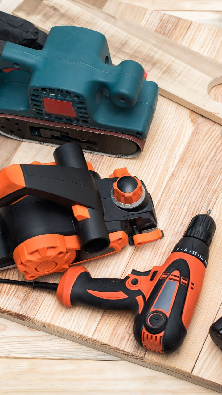 Trois outils électriques sur une surface en bois : une ponceuse à bande, une ponceuse orbitale et une perceuse