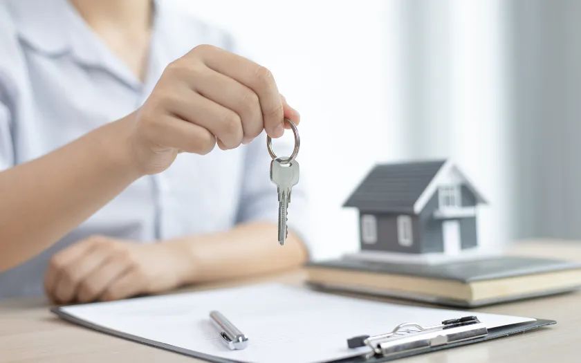Persona con llaves frente a una casa en miniatura, lo que simboliza una transacción inmobiliaria. Hay un documento y un bolígrafo sobre el escritorio.