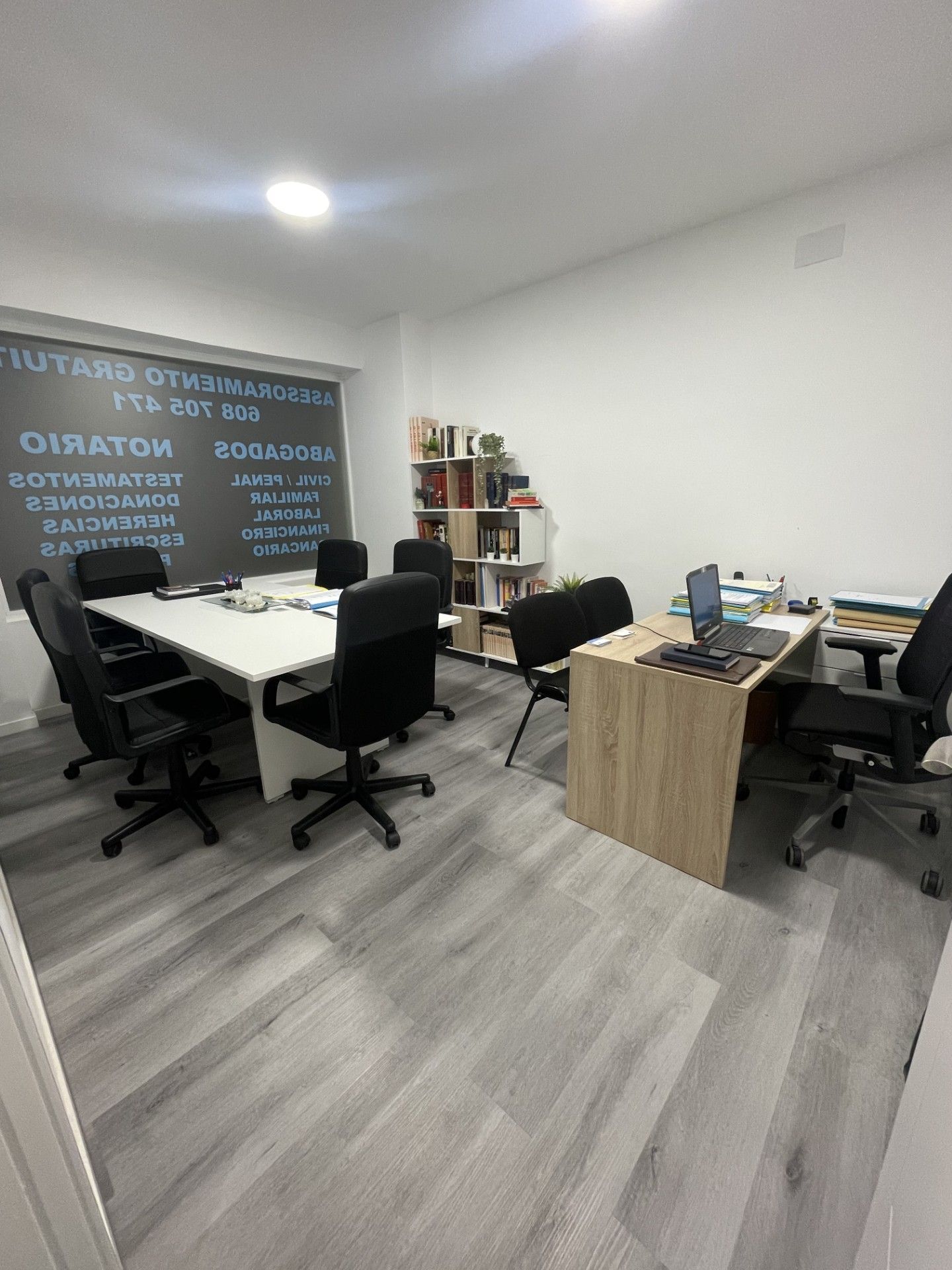 Una oficina con mesa de conferencias, escritorio y estantería. Suelo gris y paredes blancas con una ventana con texto.