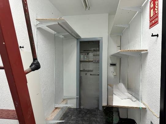 Un trastero estrecho con estantes, una puerta y una escalera a la izquierda. Paredes y estantes blancos, suelo oscuro.