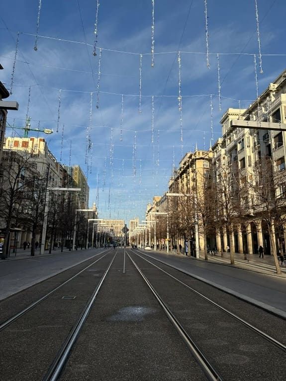 Vista de la calle, edificios a ambos lados, vías de tranvía en el centro, cielo azul, luces en lo alto.