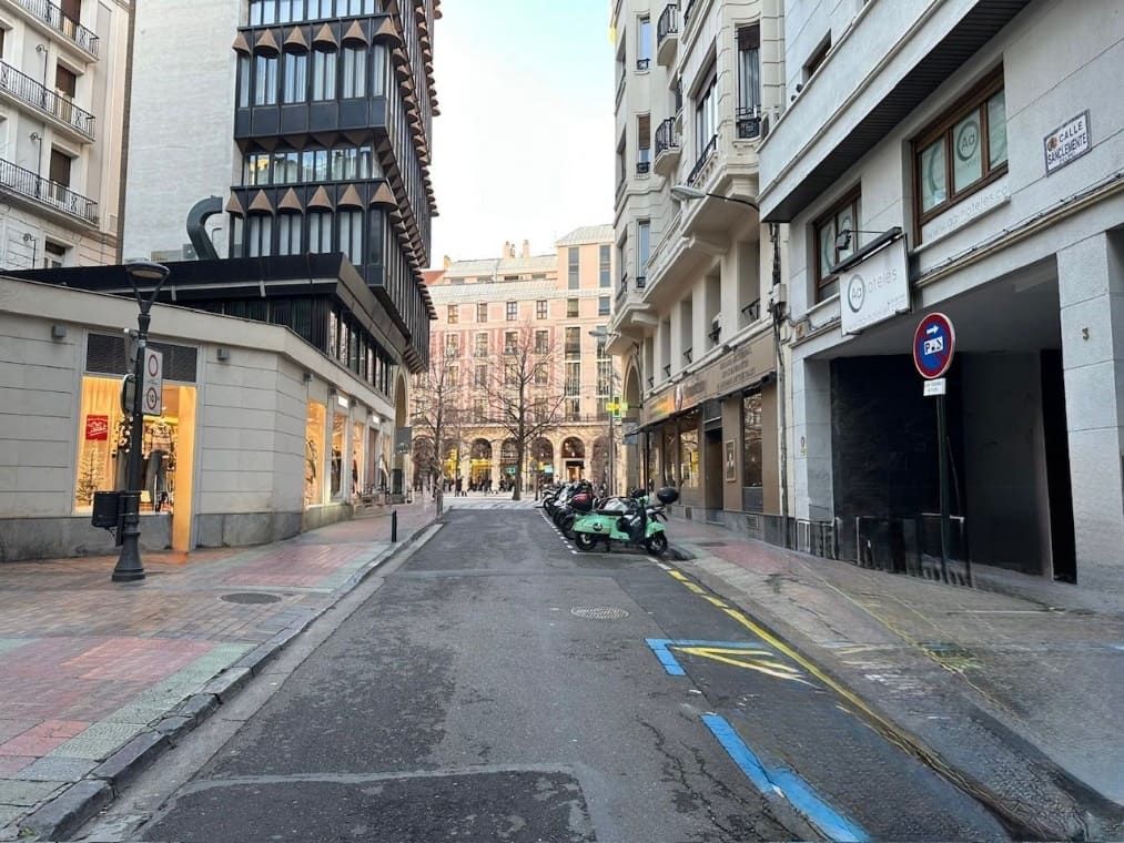 Escena callejera, edificios con escaparates a ambos lados, motos aparcadas, carretera vacía, soleado.