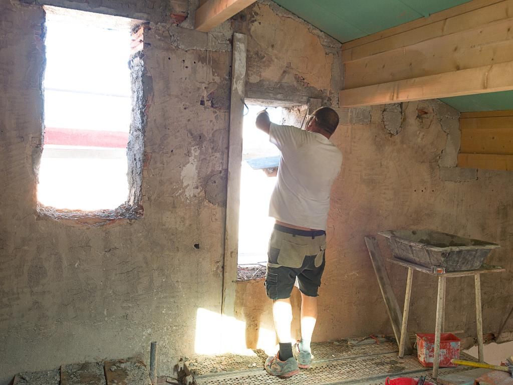 Un ouvrier enduit un mur près d'une fenêtre dans une pièce inachevée aux poutres apparentes.