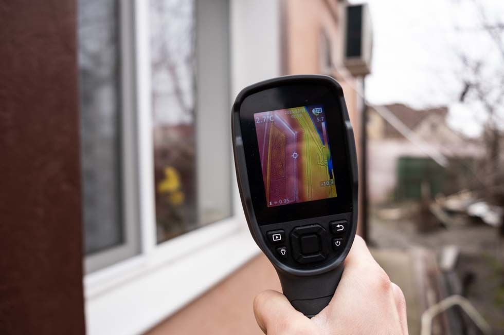 Caméra thermique portable pointée vers une fenêtre de maison, affichant des motifs thermiques sur son écran