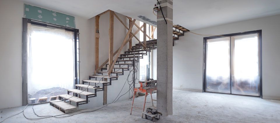 Intérieur inachevé avec escalier apparent et grandes fenêtres laissant entrer la lumière du jour.