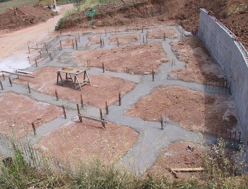 Fondations de chantier avec semelles en béton et armatures métalliques sur un terrain en terre battue, bordées par un mur de soutènement gris.