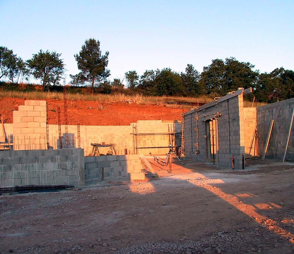 Maison en parpaings partiellement construite sur un terrain vague au coucher du soleil, avec échafaudages et flanc de colline en terre rouge en arrière-plan