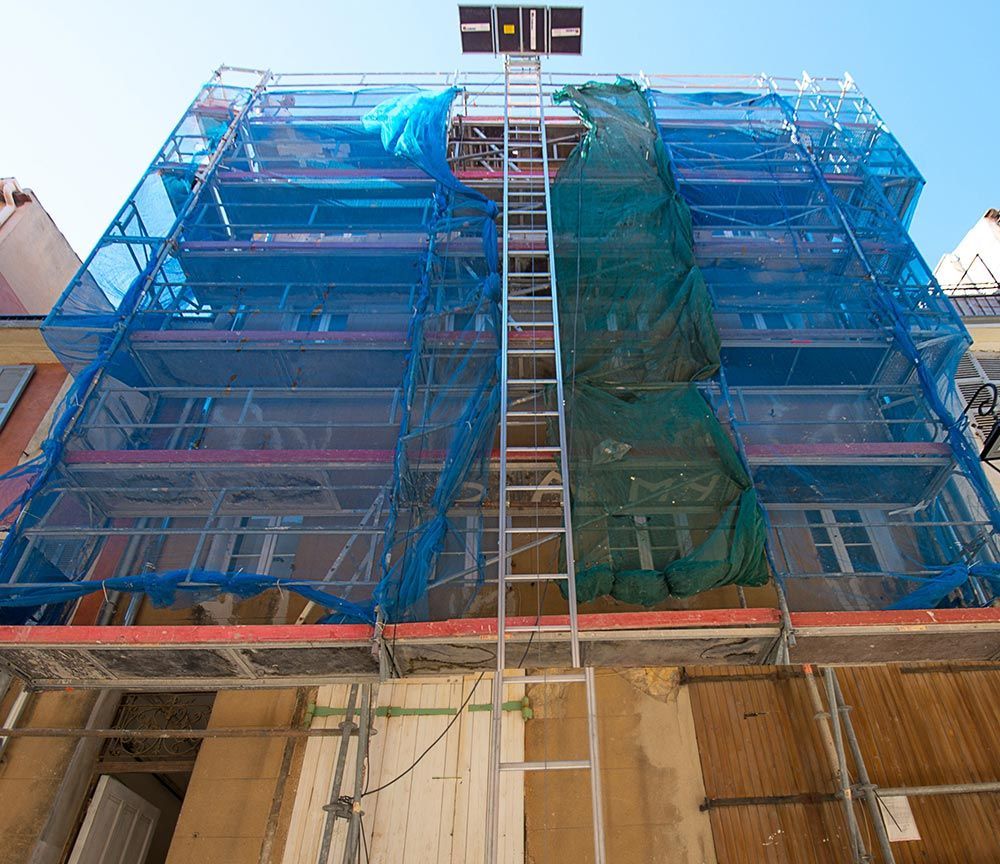 Un filet de chantier bleu recouvre la façade d'un immeuble de plusieurs étages où se trouvent des échafaudages et une échelle centrale.