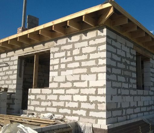 Maison en briques partiellement construite, charpente de toit inachevée, sous un ciel bleu clair.
