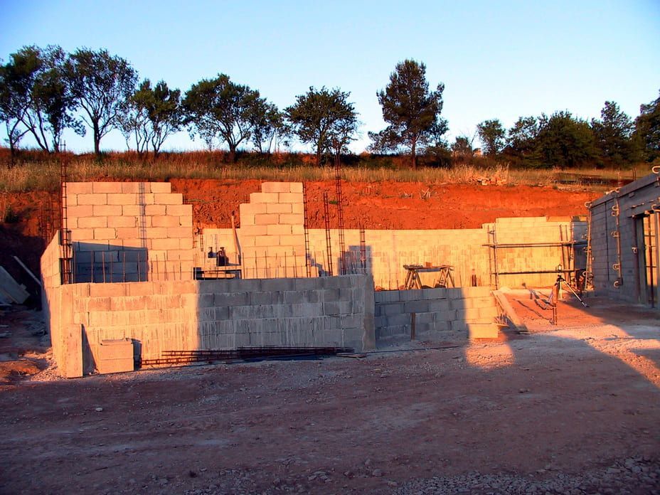 Construction en parpaings partiellement édifiée sur un terrain vague au coucher du soleil, avec des arbres et un ciel bleu en arrière-plan.