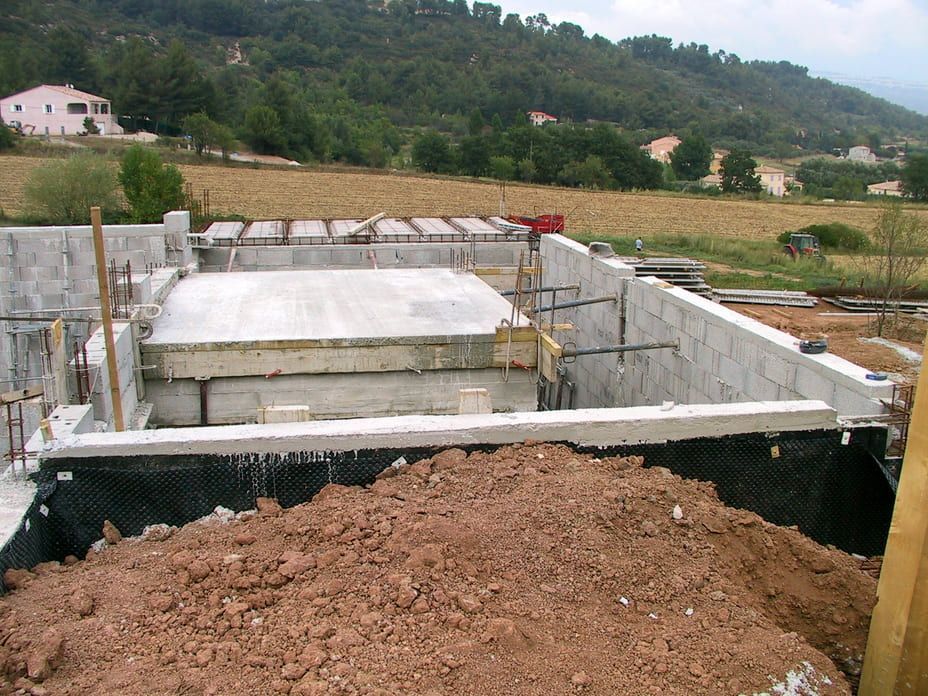 Fondations en béton partiellement construites sur un flanc de colline rurale, avec un premier plan de terre et des champs à l'arrière-plan.