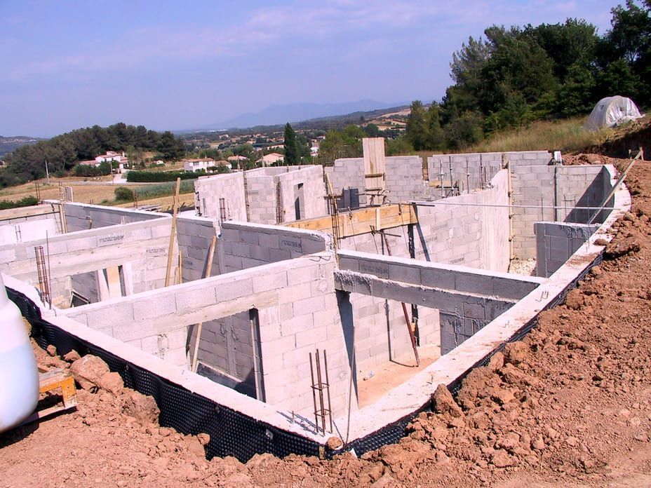 Murs de fondation en béton d'une maison en construction sur un terrain en terre battue, avec des montagnes en arrière-plan.
