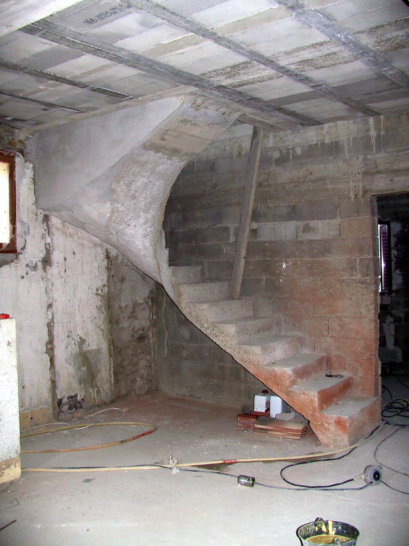 Escalier en béton brut à l'intérieur d'une pièce nue aux murs apparents et aux fils électriques éparpillés.