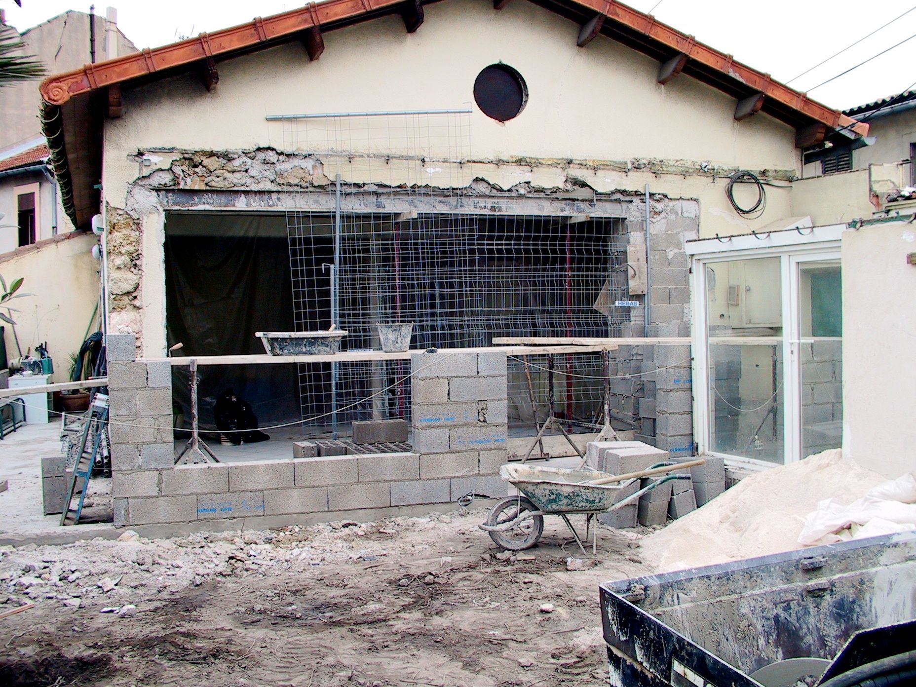 Maison partiellement démolie, murs apparents, gravats et matériaux de construction devant.