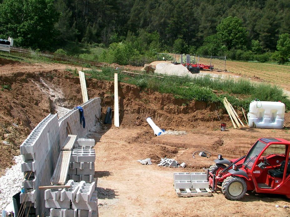 Chantier de construction avec des parpaings empilés, une tranchée de terre et un chariot utilitaire rouge à l'extérieur.