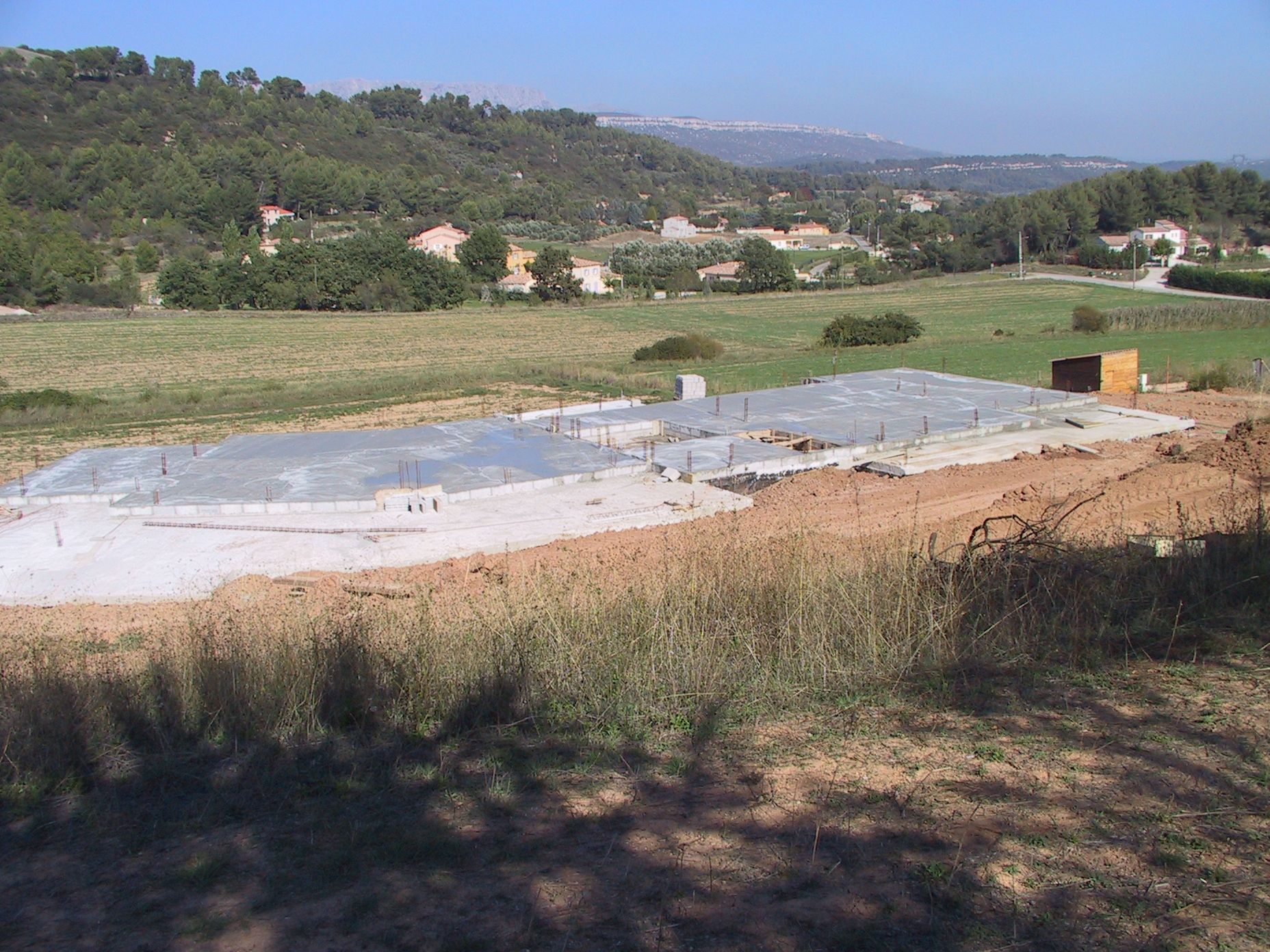 Fondations en béton dans un champ herbeux, avec des collines et des maisons en arrière-plan, sous un ciel dégagé.