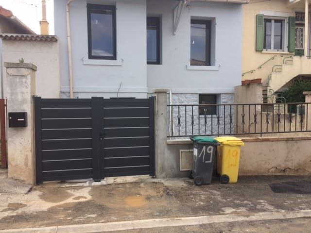 Entrée de la maison avec portail sombre, petit muret en façade et deux poubelles sur le trottoir.