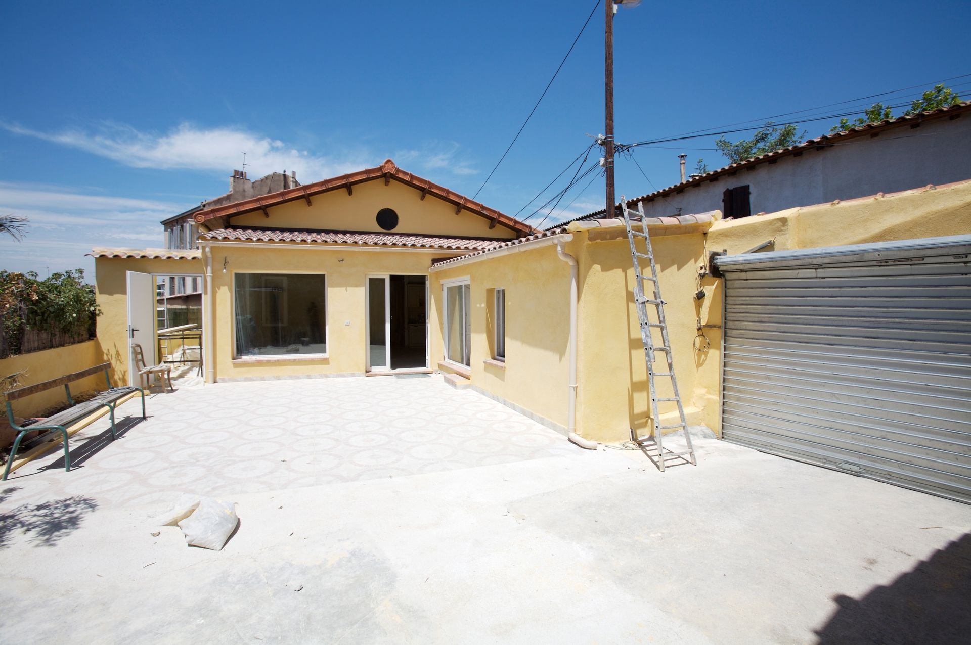 Cour ensoleillée d'une petite maison aux murs jaunes, au sol blanc et à la porte de garage métallique, sous un ciel bleu.