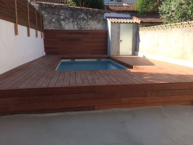 Petite terrasse dans le jardin avec piscine carrée, murs en bois et façades en stuc blanc baignées de soleil.