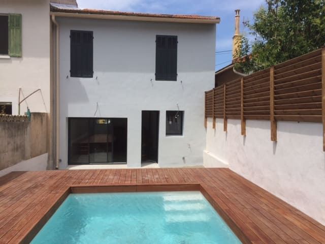 Jardin avec une petite piscine turquoise, une terrasse en bois et une maison blanche à deux étages avec des volets foncés.