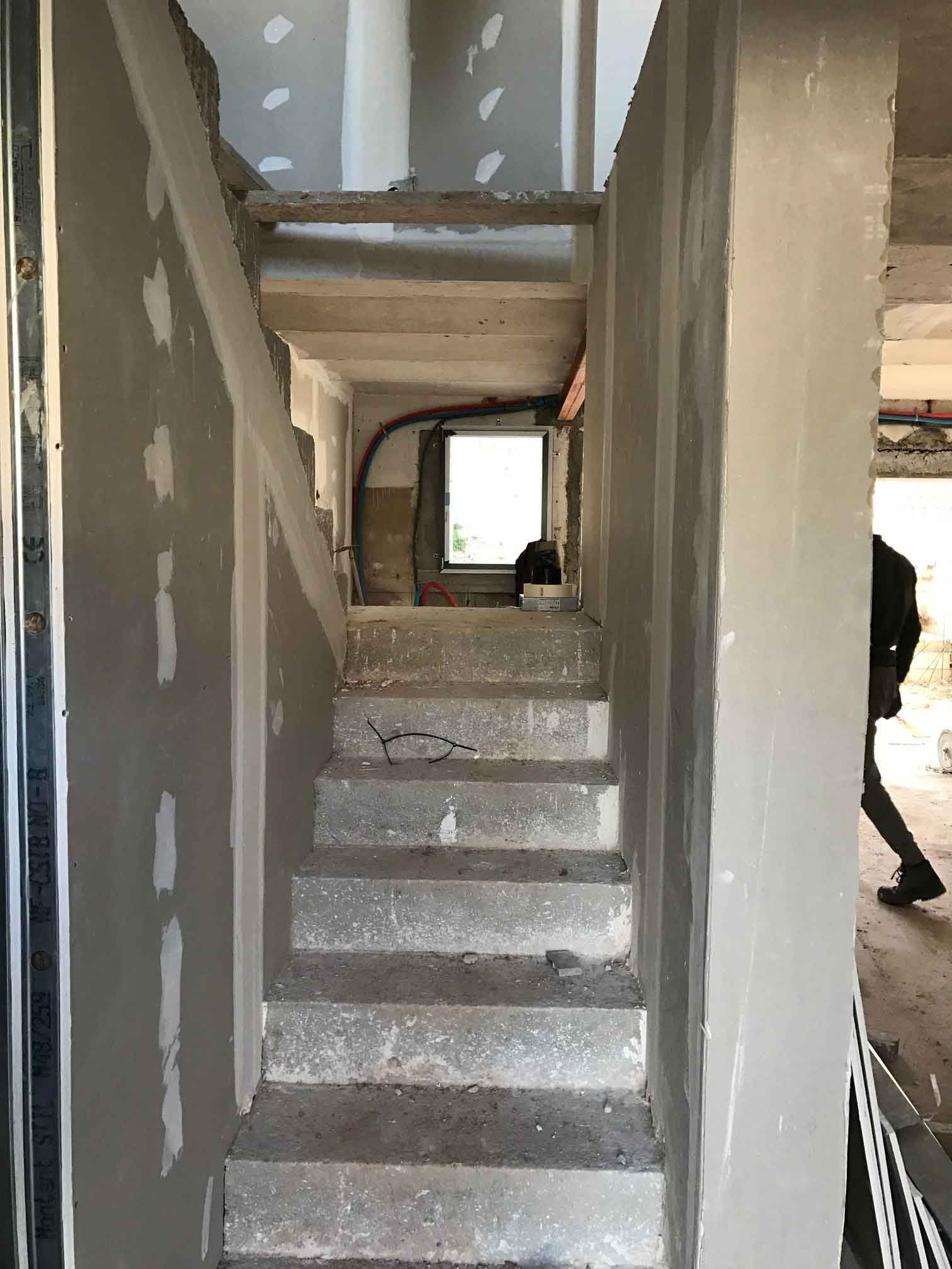 Escalier étroit et inachevé à l'intérieur d'un bâtiment aux murs en plâtre gris et aux poutres apparentes au plafond.