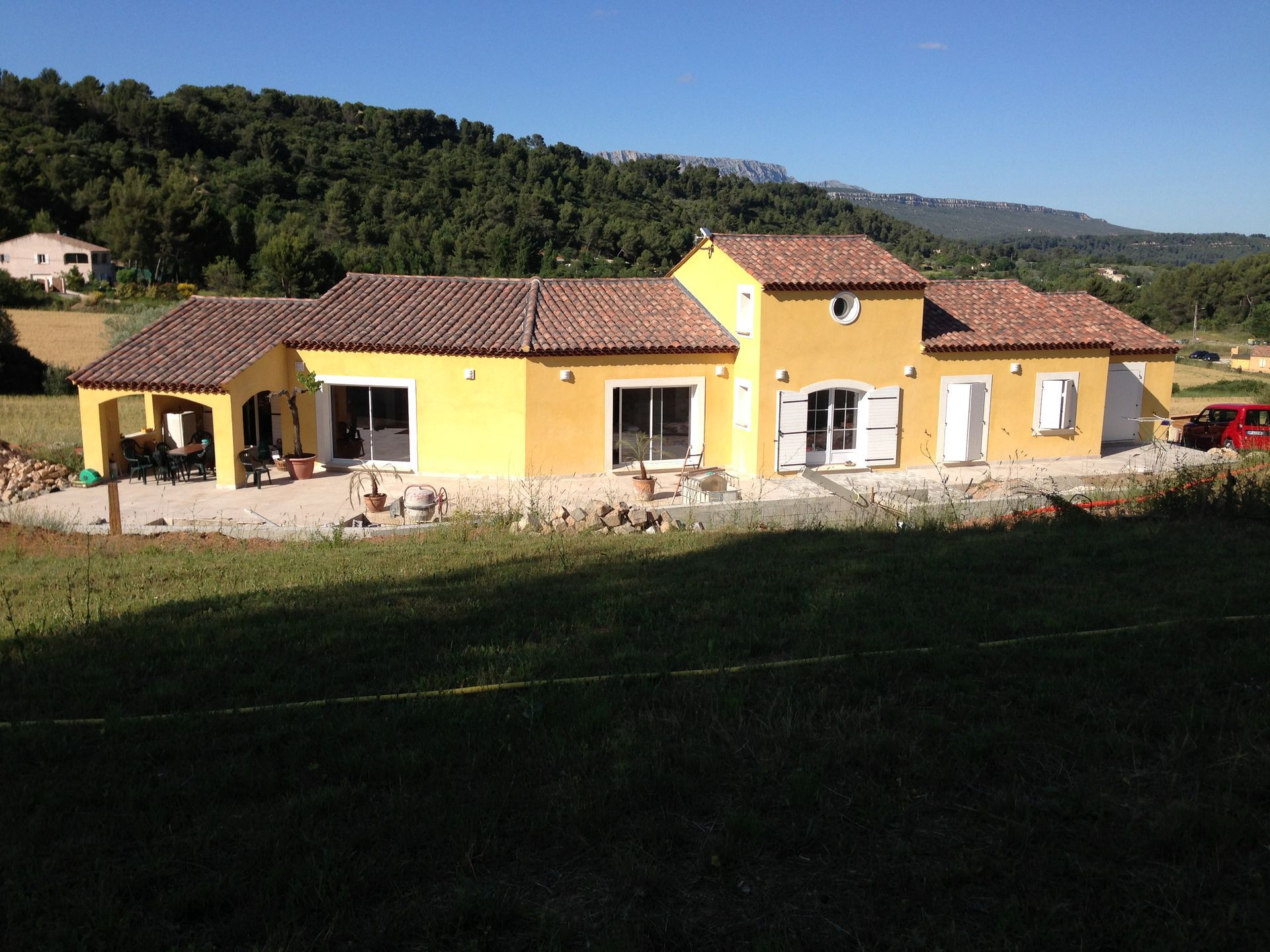 Maison rurale jaune avec un toit de tuiles rouges et un petit porche couvert dans un cadre ensoleillé à flanc de colline