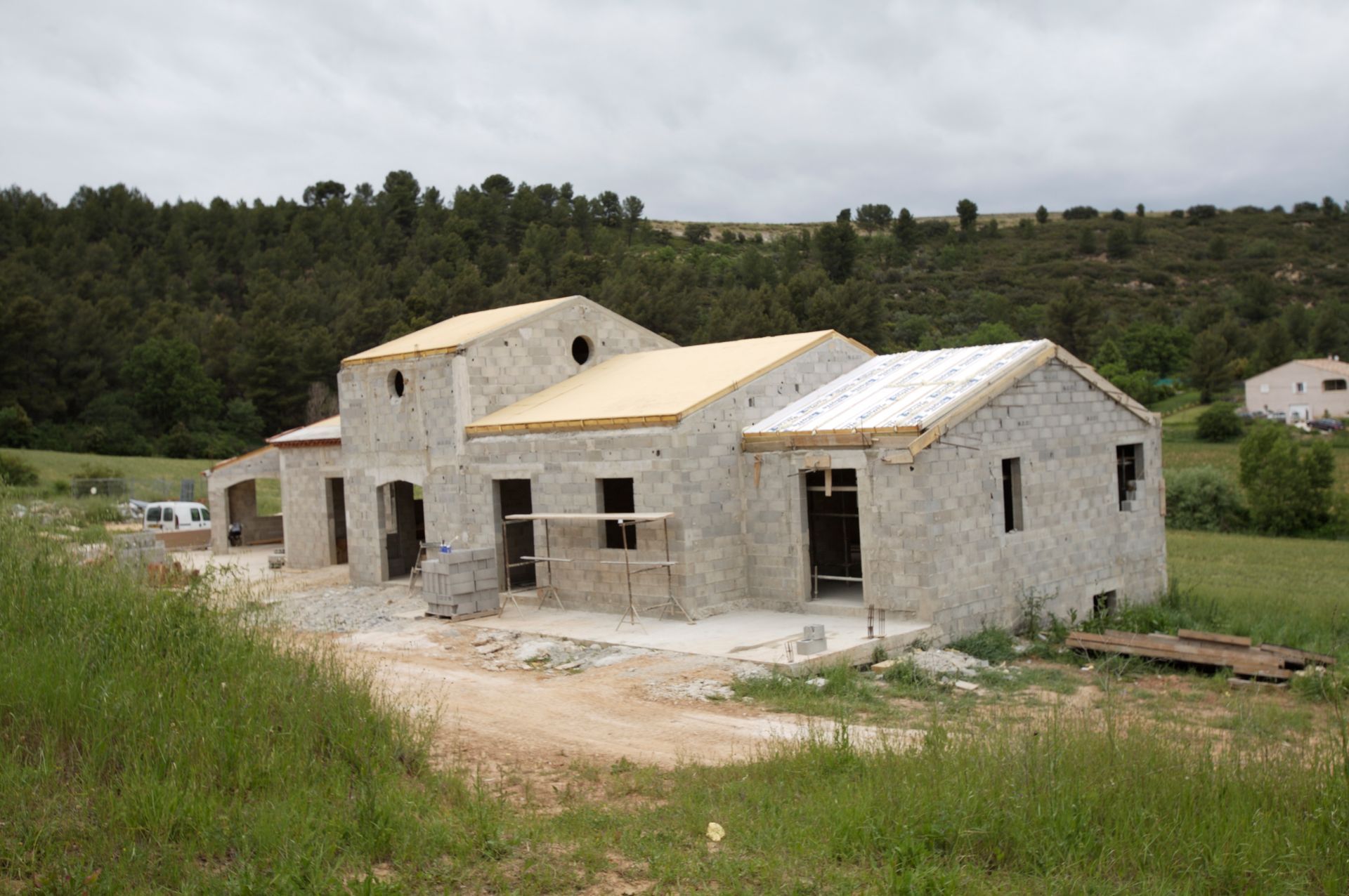 Maison en pierre en construction dans un champ, avec des collines verdoyantes et des arbres en arrière-plan.