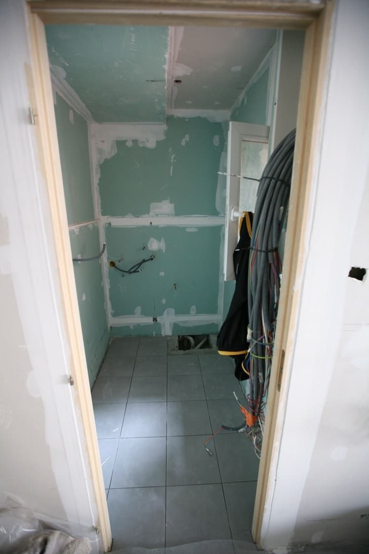 Petit placard ou buanderie inachevé avec cloisons sèches vertes, sol carrelé et vêtements suspendus à l'intérieur d'une porte ouverte.