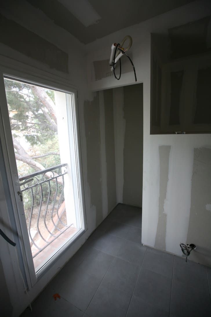 Petite pièce inachevée aux murs gris, sol carrelé, porte-fenêtre donnant sur un balcon et câblage apparent au plafond.