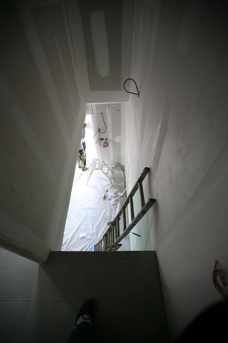Vue plongeante sur un escalier étroit menant à une porte lumineuse, avec une rampe sur la droite.